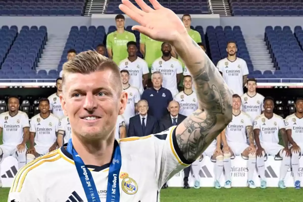 Un jugador de fútbol con uniforme blanco y una medalla en el cuello saluda frente a un grupo de personas, posiblemente su equipo, que están sentados y de pie en un estadio vacío.