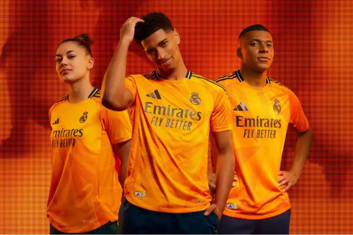 Tres personas posando con la camiseta naranja del Real Madrid con el logo de Emirates Fly Better y un fondo naranja.