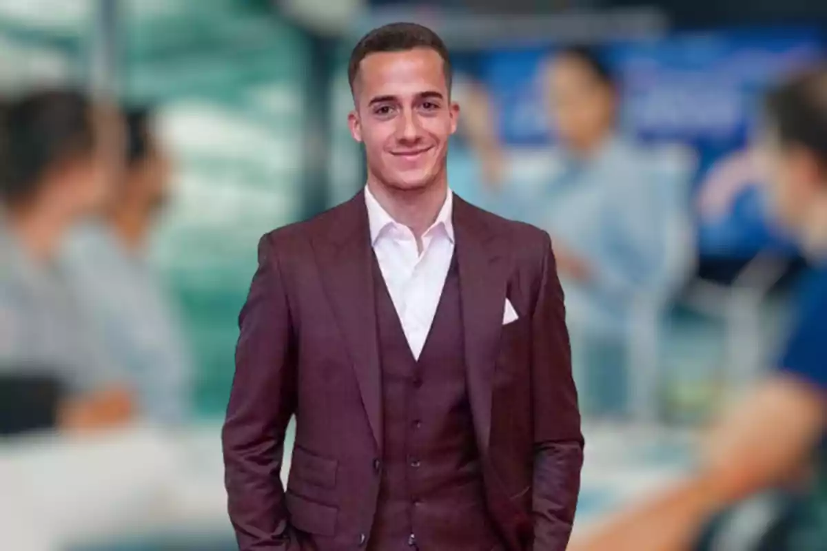 Lucas Vázquez con traje marrón y camisa blanca, sonriendo, con un fondo borroso de personas en una oficina.