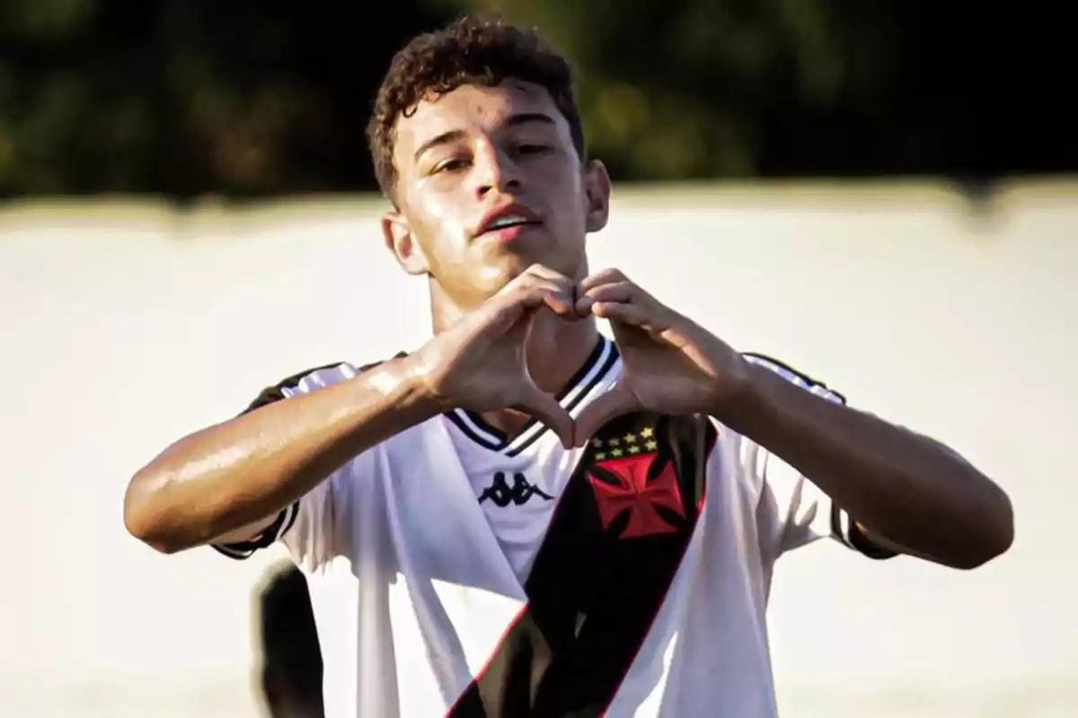 Joven futbolista haciendo un gesto de corazón con las manos mientras viste una camiseta de fútbol blanca con una franja diagonal negra y roja.