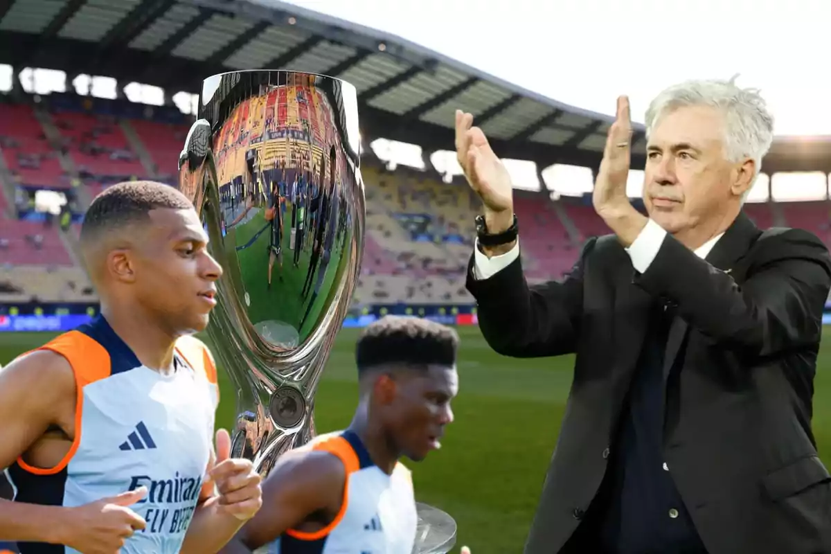 Jugadores de fútbol y un entrenador aplaudiendo junto a un trofeo en un estadio.