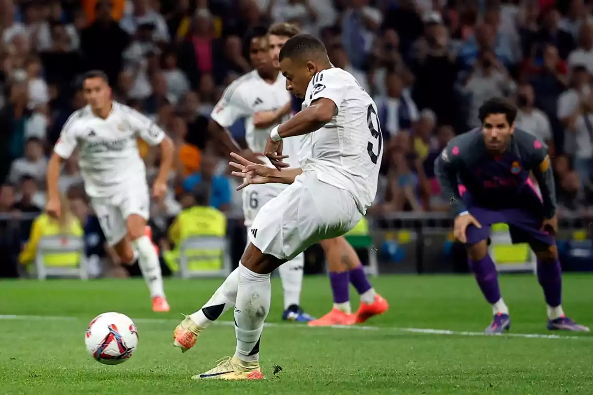 Un jugador de fútbol en uniforme blanco pateando un balón durante un partido mientras otros jugadores y el portero observan.