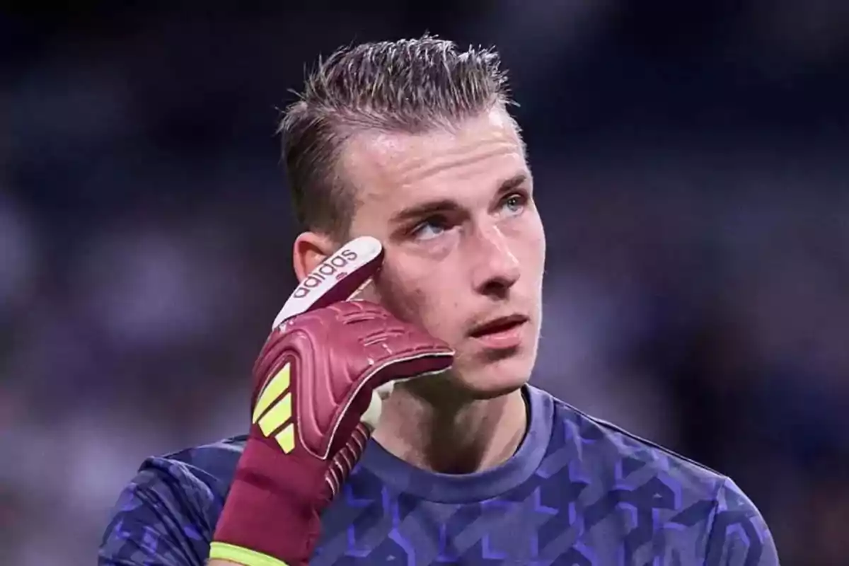 Un portero de fútbol con guantes rojos y camiseta azul oscuro se toca la sien con una expresión pensativa.