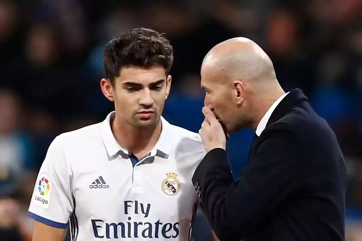 Un jugador de fútbol con la camiseta del Real Madrid escucha atentamente a un hombre calvo que le habla al oído.