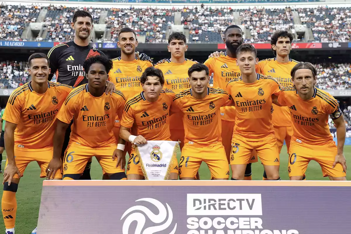 Jugadores del Real Madrid posando para una foto de equipo en el campo de fútbol, vistiendo uniformes naranjas y sonriendo frente a una multitud de espectadores.