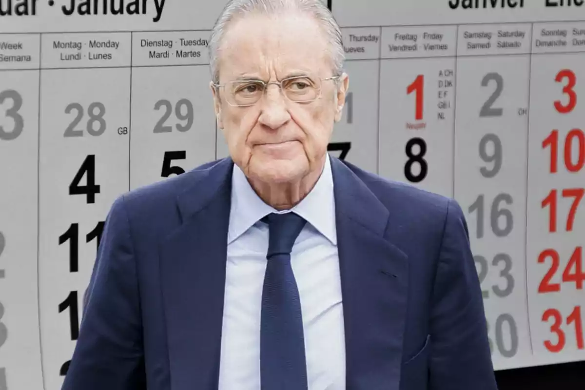 Un hombre mayor con gafas y traje azul oscuro está de pie frente a un calendario grande que muestra el mes de enero.