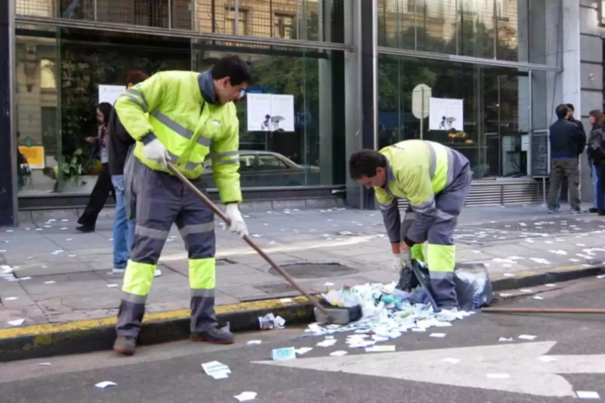 Dos trabajadores de limpieza con uniformes reflectantes recogen papeles en una acera frente a un edificio de vidrio.