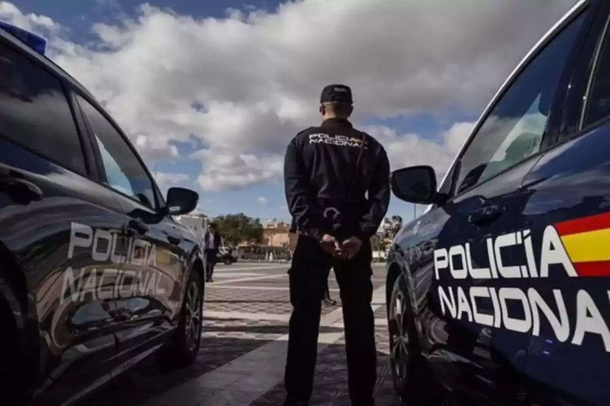 Un agente de la Policía Nacional de espaldas entre dos coches patrulla en un día soleado con nubes. Un agente de la Policía Nacional de espaldas entre dos coches patrulla en un día soleado con nubes.