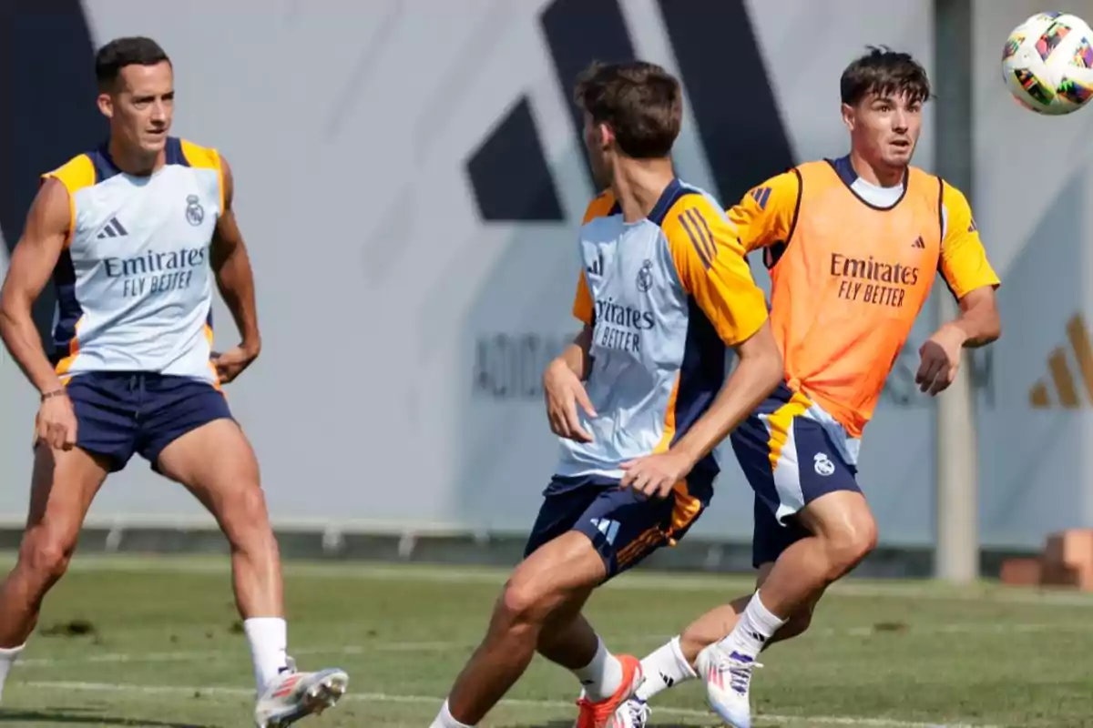 Jugadores de fútbol del Real Madrid entrenando en el campo, uno de ellos con un peto naranja y los otros dos con camisetas de entrenamiento blancas y azules.