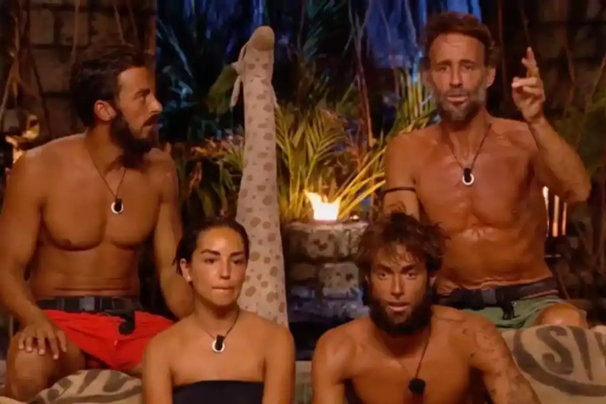 Cuatro personas con collares negros están sentadas en un entorno selvático, una pierna con calcetín de jirafa se eleva en el centro de la imagen, todos lucen ropa de playa y parecen estar participando en un reality show. Cuatro personas con collares negros están sentadas en un entorno selvático, una pierna con calcetín de jirafa se eleva en el centro de la imagen, todos lucen ropa de playa y parecen estar participando en un reality show.