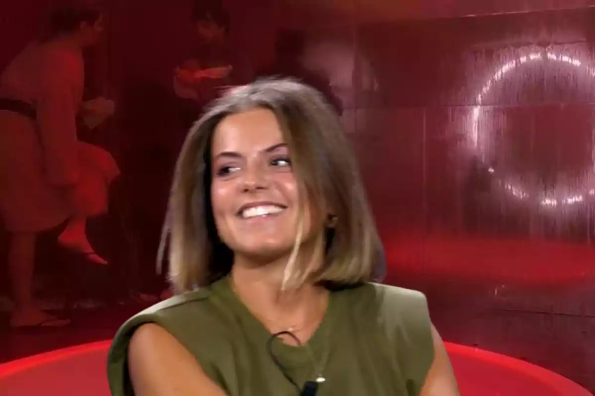 Una mujer sonriente con cabello corto y rubio oscuro, vestida con una camiseta verde, en un entorno con iluminación roja y figuras borrosas en el fondo.