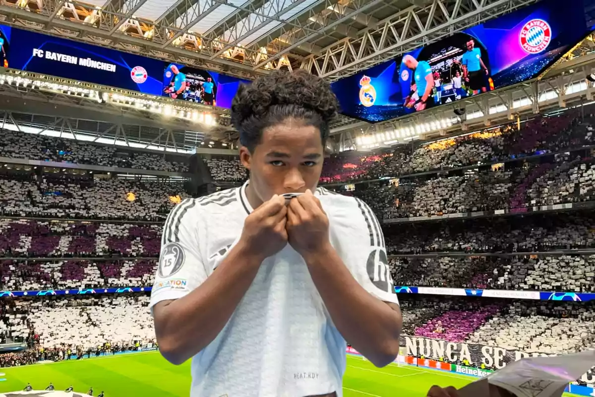 Un jugador de fútbol con una camiseta blanca besa el escudo en un estadio lleno de aficionados, con pantallas que muestran el logo del FC Bayern München y el Real Madrid.