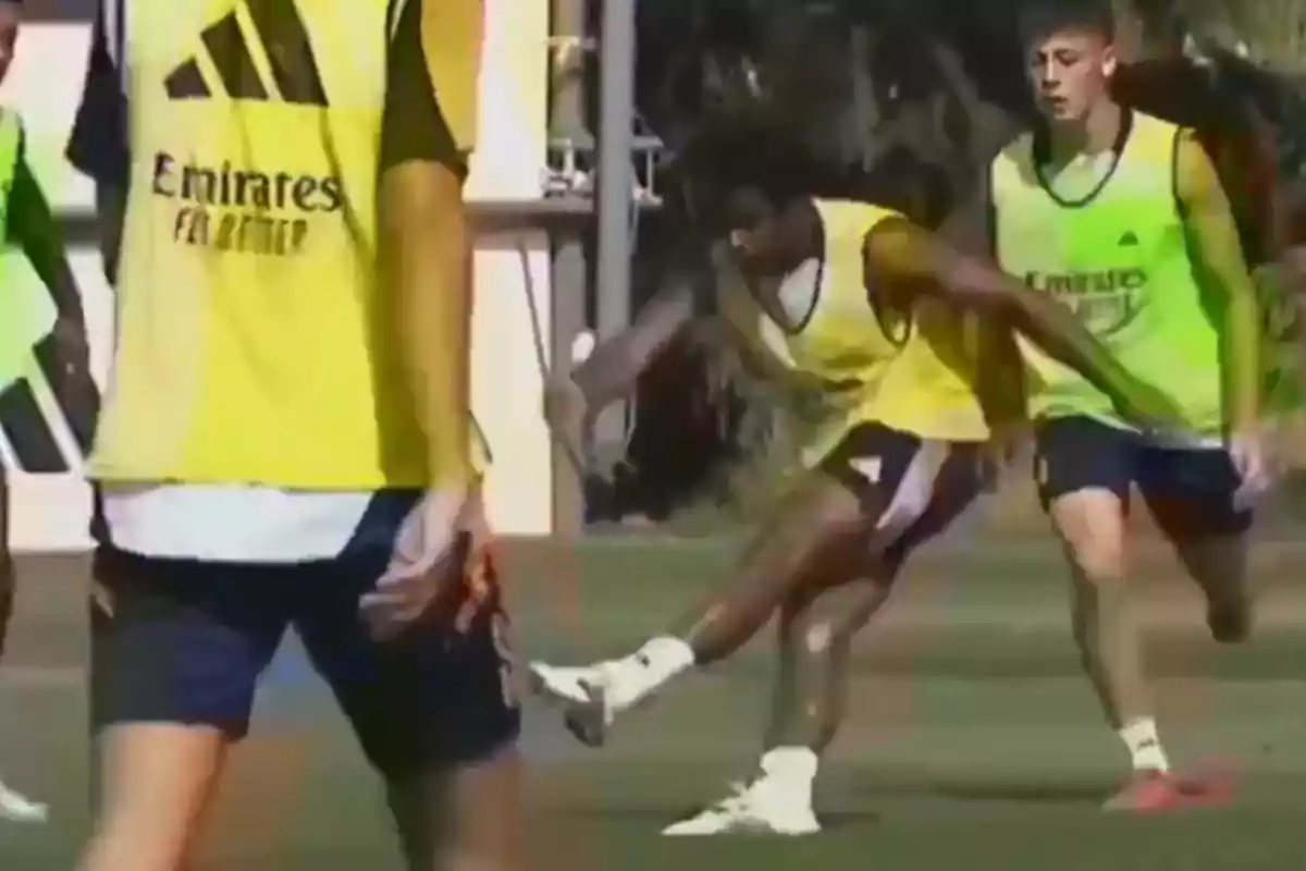 Jugadores de fútbol entrenando en un campo, usando chalecos amarillos y uniformes deportivos.