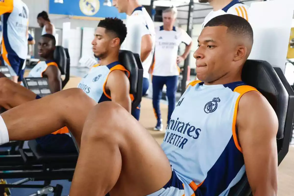 Jugadores de fútbol entrenando en el gimnasio con camisetas de su equipo mientras realizan ejercicios de piernas en máquinas de pesas.