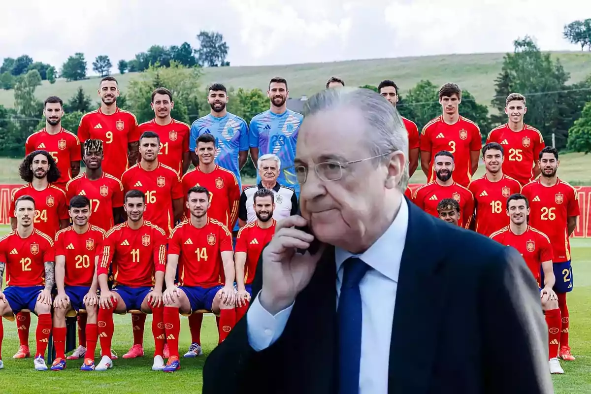 Un grupo de jugadores de fútbol con uniformes rojos posando para una foto de equipo en un campo, con un hombre mayor en traje hablando por teléfono en primer plano.