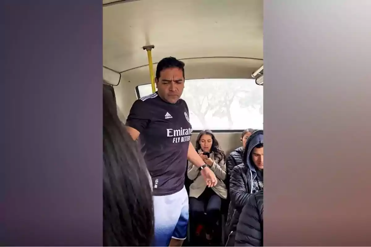 Un hombre de pie en un transporte público, usando una camiseta deportiva negra, mientras otras personas están sentadas y usando sus teléfonos.