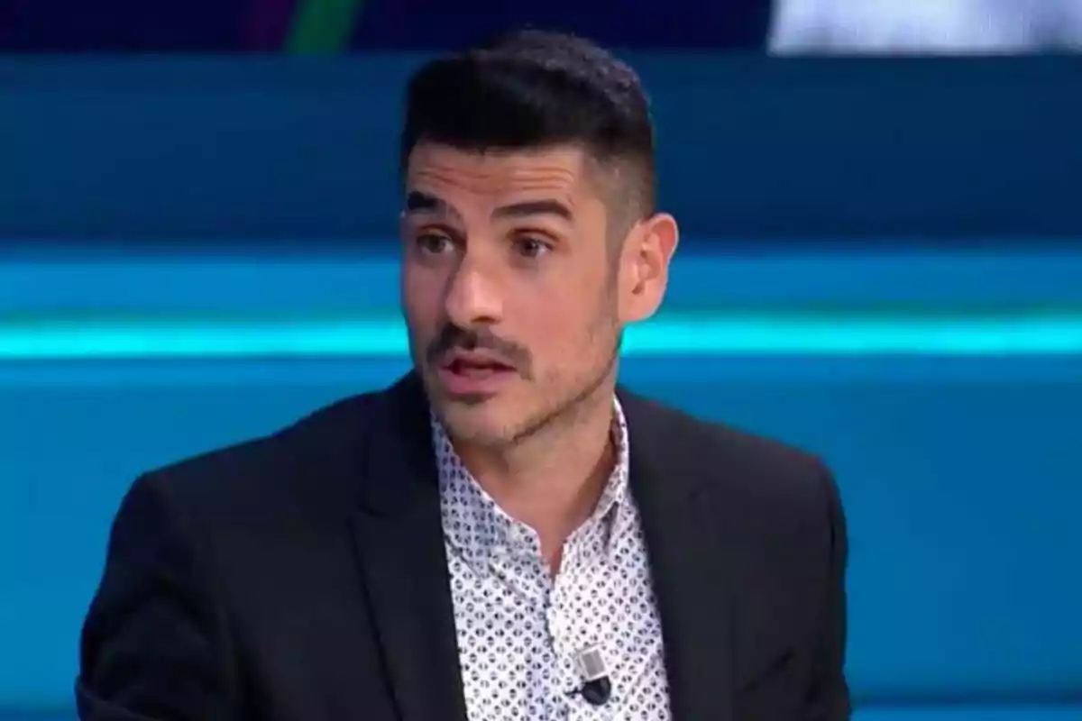 Hombre con barba y bigote, vestido con camisa blanca estampada y saco negro, hablando en un programa de televisión.