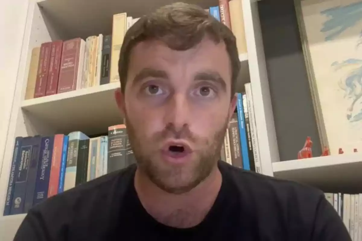 Un hombre con barba y cabello corto está hablando frente a una estantería llena de libros.