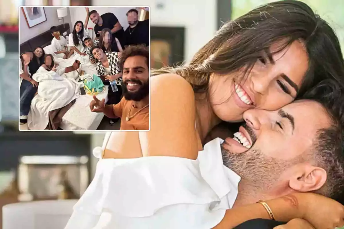 Una pareja sonriente se abraza mientras un grupo de personas posa para una selfie en una habitación.