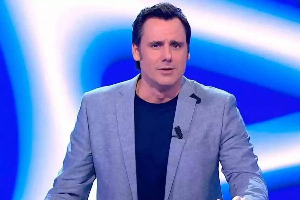Un hombre con chaqueta gris y camiseta oscura está de pie frente a un fondo azul y blanco. Un hombre con chaqueta gris y camiseta oscura está de pie frente a un fondo azul y blanco.