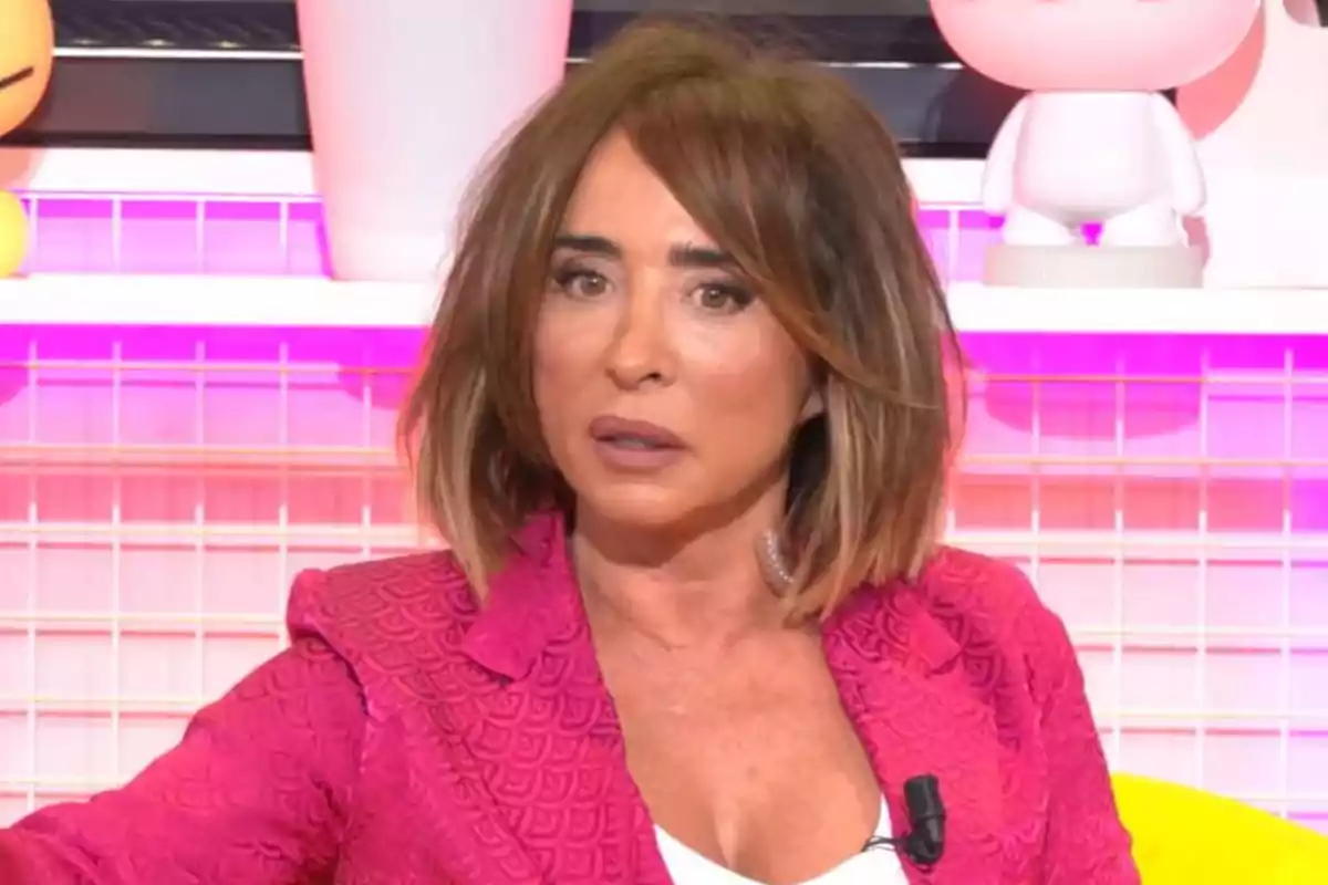 Una persona con cabello castaño y un atuendo rosa está sentada en un entorno colorido con luces de neón y figuras decorativas en el fondo. Una persona con cabello castaño y un atuendo rosa está sentada en un entorno colorido con luces de neón y figuras decorativas en el fondo.