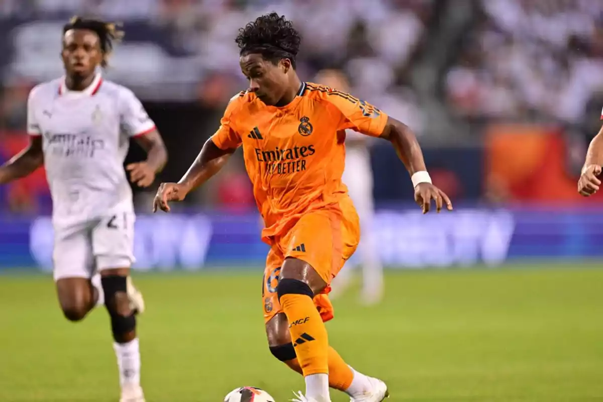 Un jugador de fútbol con uniforme naranja del Real Madrid controla el balón mientras es perseguido por un jugador del equipo contrario con uniforme blanco durante un partido.