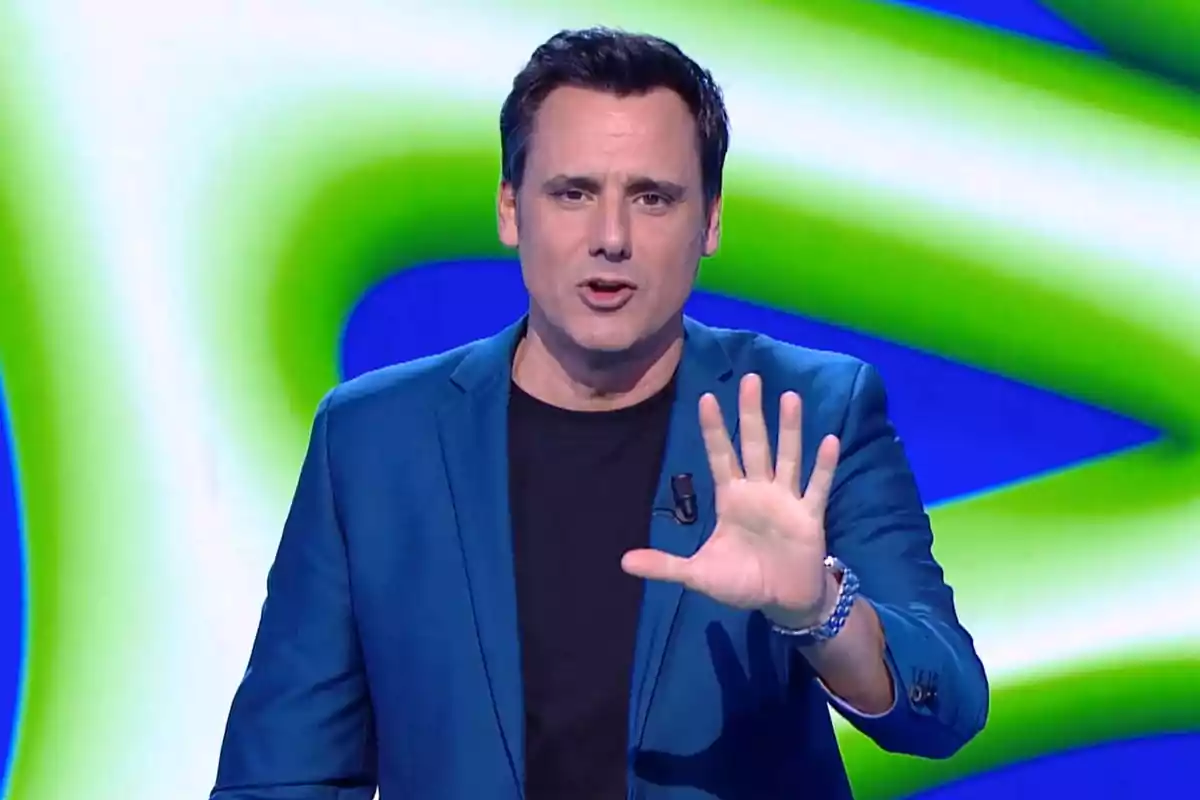Hombre con chaqueta azul gesticulando con la mano levantada frente a un fondo colorido. Hombre con chaqueta azul gesticulando con la mano levantada frente a un fondo colorido.