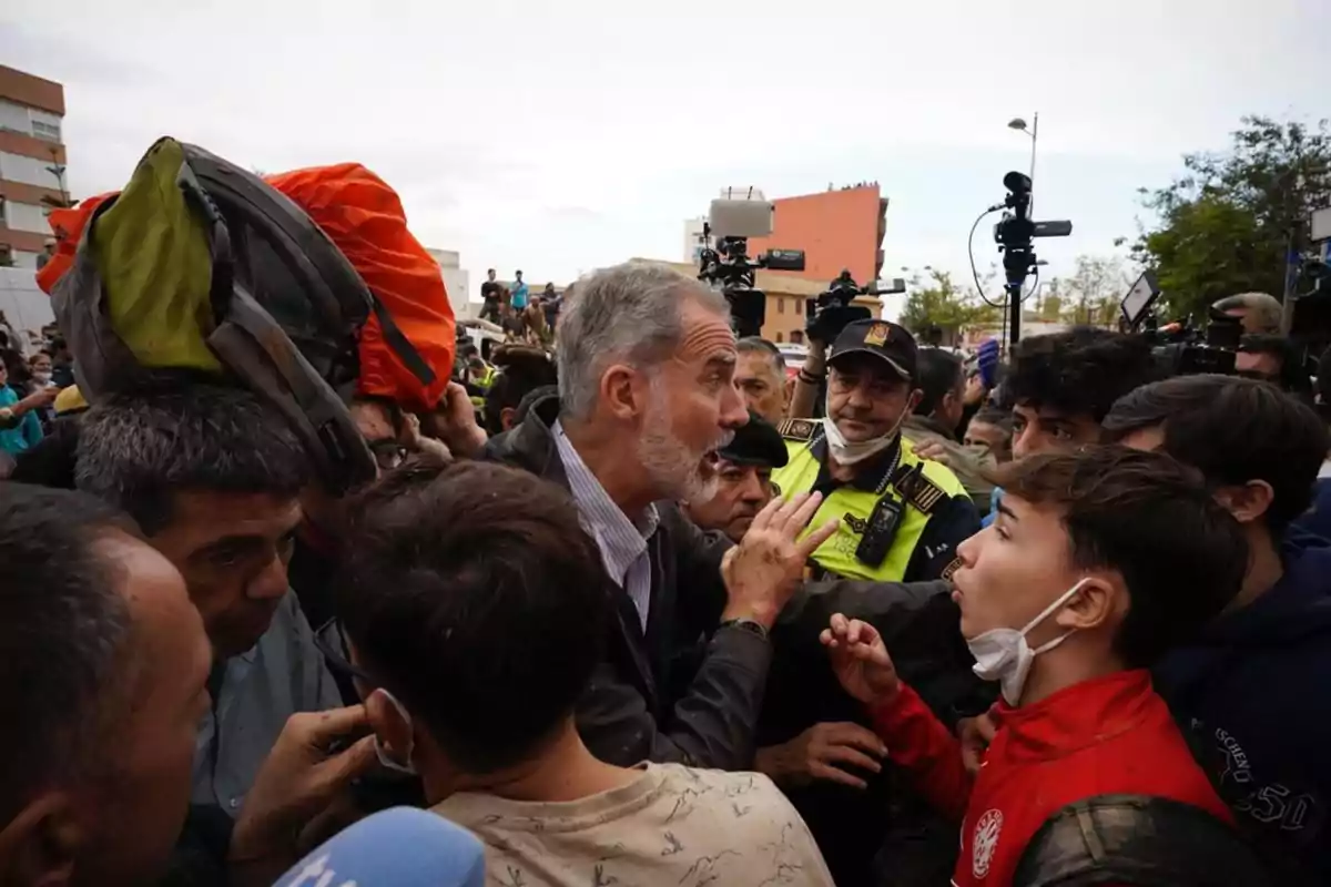 Un grupo de personas se reúne en una calle, algunas con mascarillas, mientras un hombre mayor gesticula y habla; hay presencia policial y cámaras de televisión en el fondo.