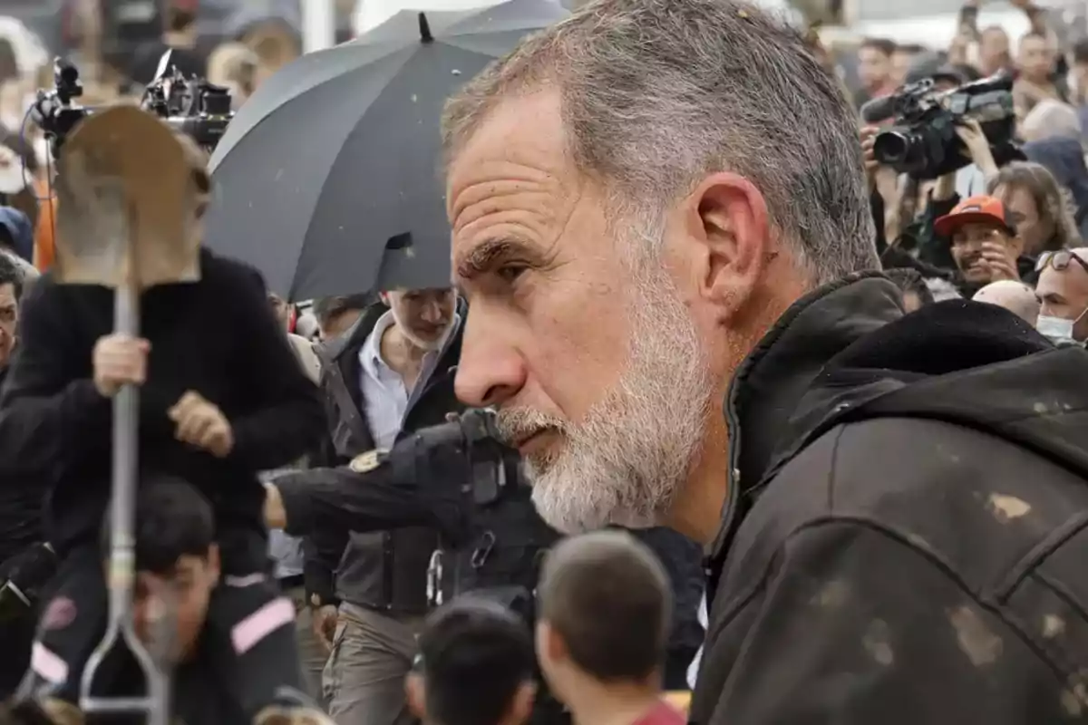 Un hombre de perfil con barba gris en un evento al aire libre rodeado de personas y cámaras.