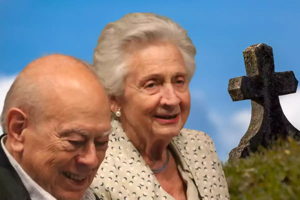 Una pareja de ancianos sonrientes junto a una cruz de piedra con un cielo azul de fondo.