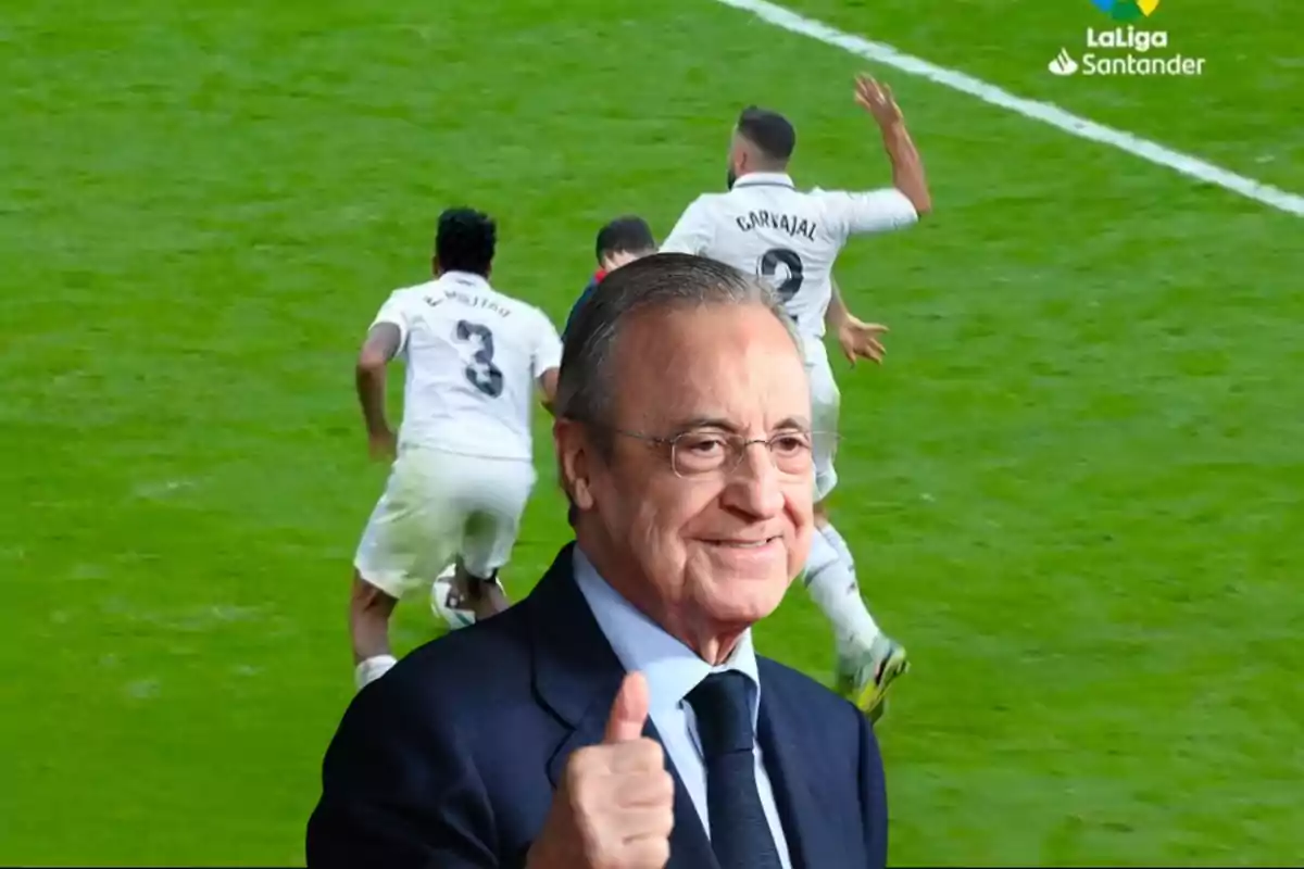 Un hombre con gafas y traje sonríe y hace un gesto de aprobación con el pulgar hacia arriba, mientras al fondo se observa un partido de fútbol de LaLiga Santander con jugadores en acción.