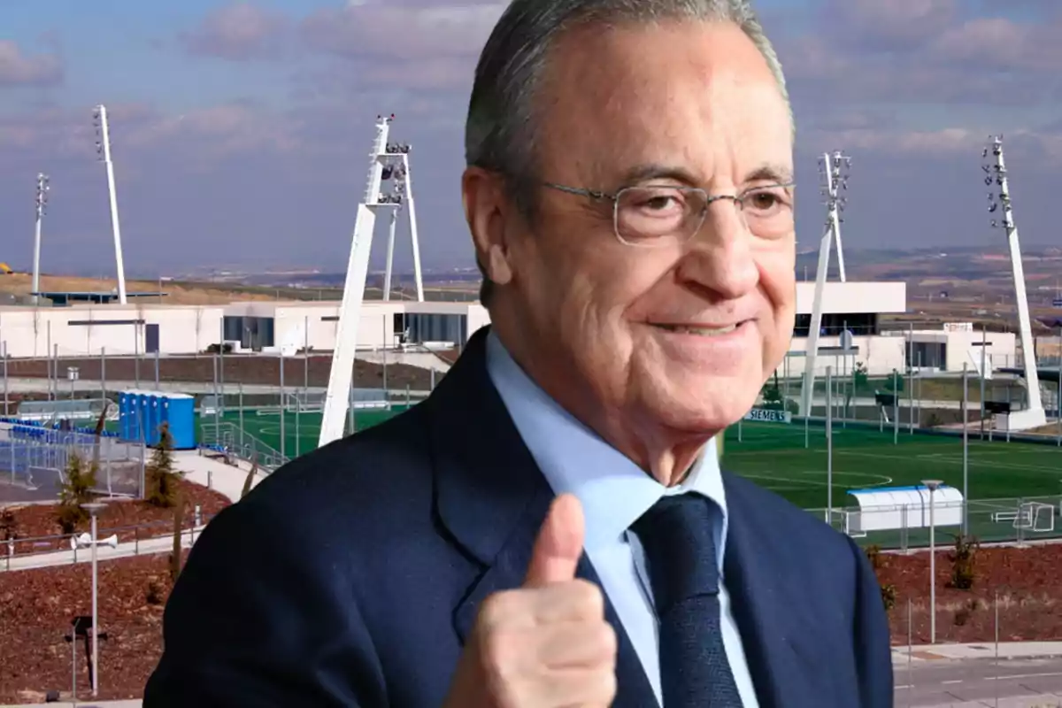 Hombre con gafas y traje oscuro haciendo un gesto de aprobación con el pulgar levantado, con un campo de entrenamiento deportivo en el fondo.