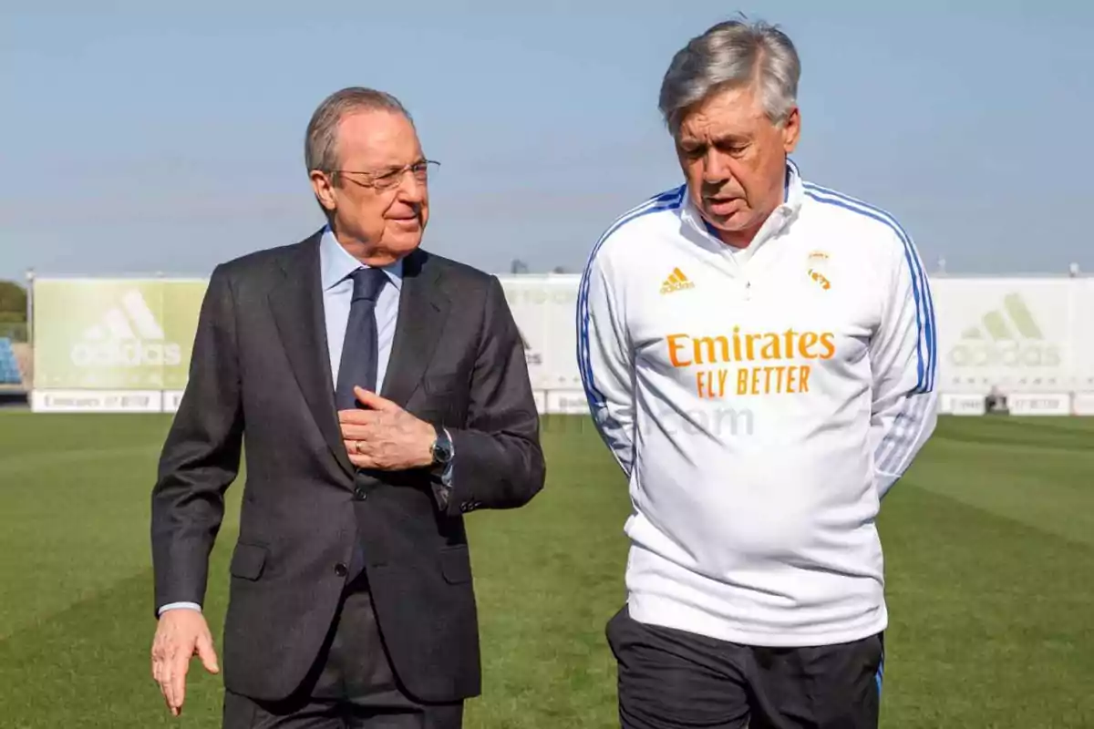 Dos hombres caminando y conversando en un campo de fútbol, uno con traje y el otro con ropa deportiva del Real Madrid.