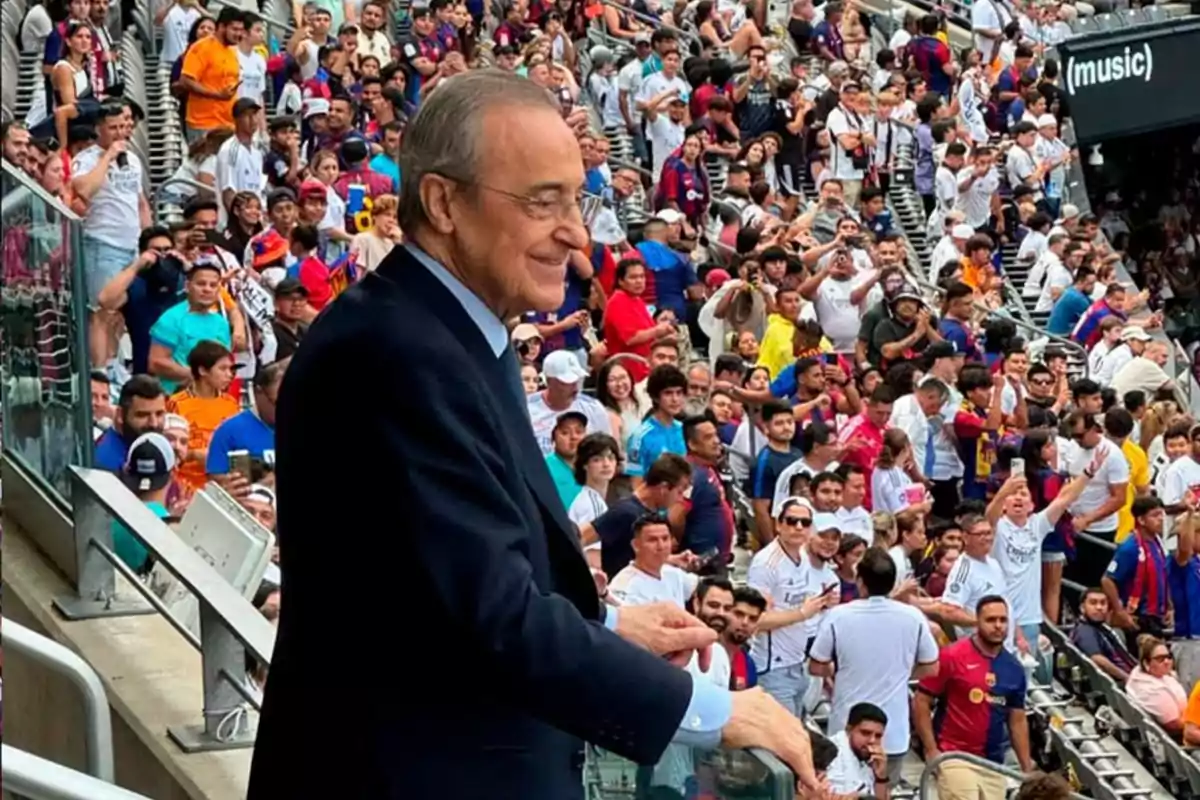 Un hombre mayor con traje oscuro sonríe mientras está de pie en un estadio lleno de aficionados.