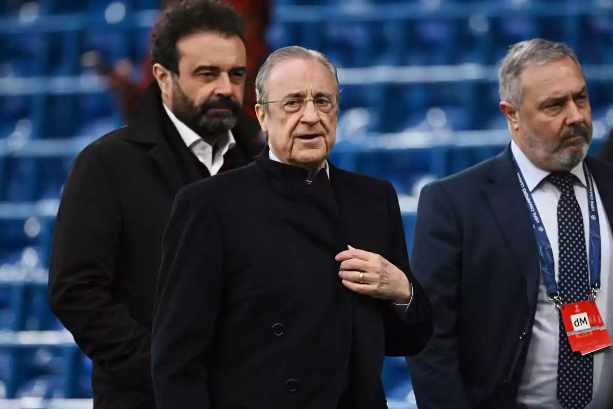 Tres hombres con trajes oscuros caminando en un estadio con asientos azules.