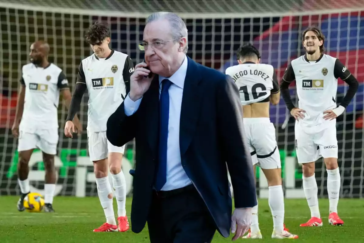 Un hombre de traje hablando por teléfono frente a un grupo de futbolistas con uniformes blancos en un campo de fútbol.