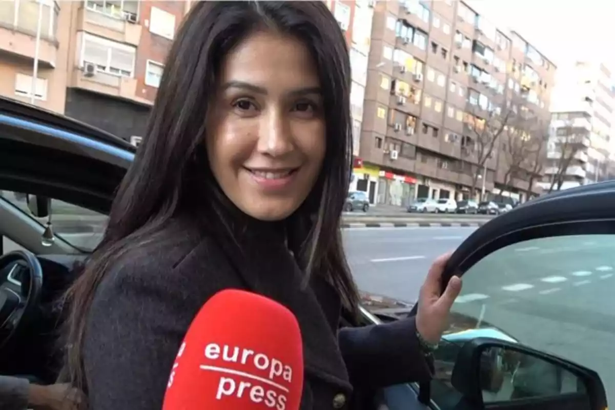Una mujer sonriente junto a un coche con un micrófono de Europa Press en primer plano en una calle urbana. Una mujer sonriente junto a un coche con un micrófono de Europa Press en primer plano en una calle urbana.