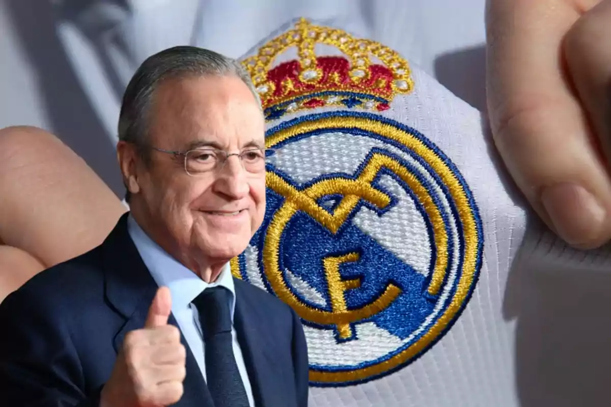 Un hombre con gafas y traje sonríe y hace un gesto de aprobación con el pulgar hacia arriba, mientras en el fondo se ve el escudo del Real Madrid bordado en una camiseta blanca.