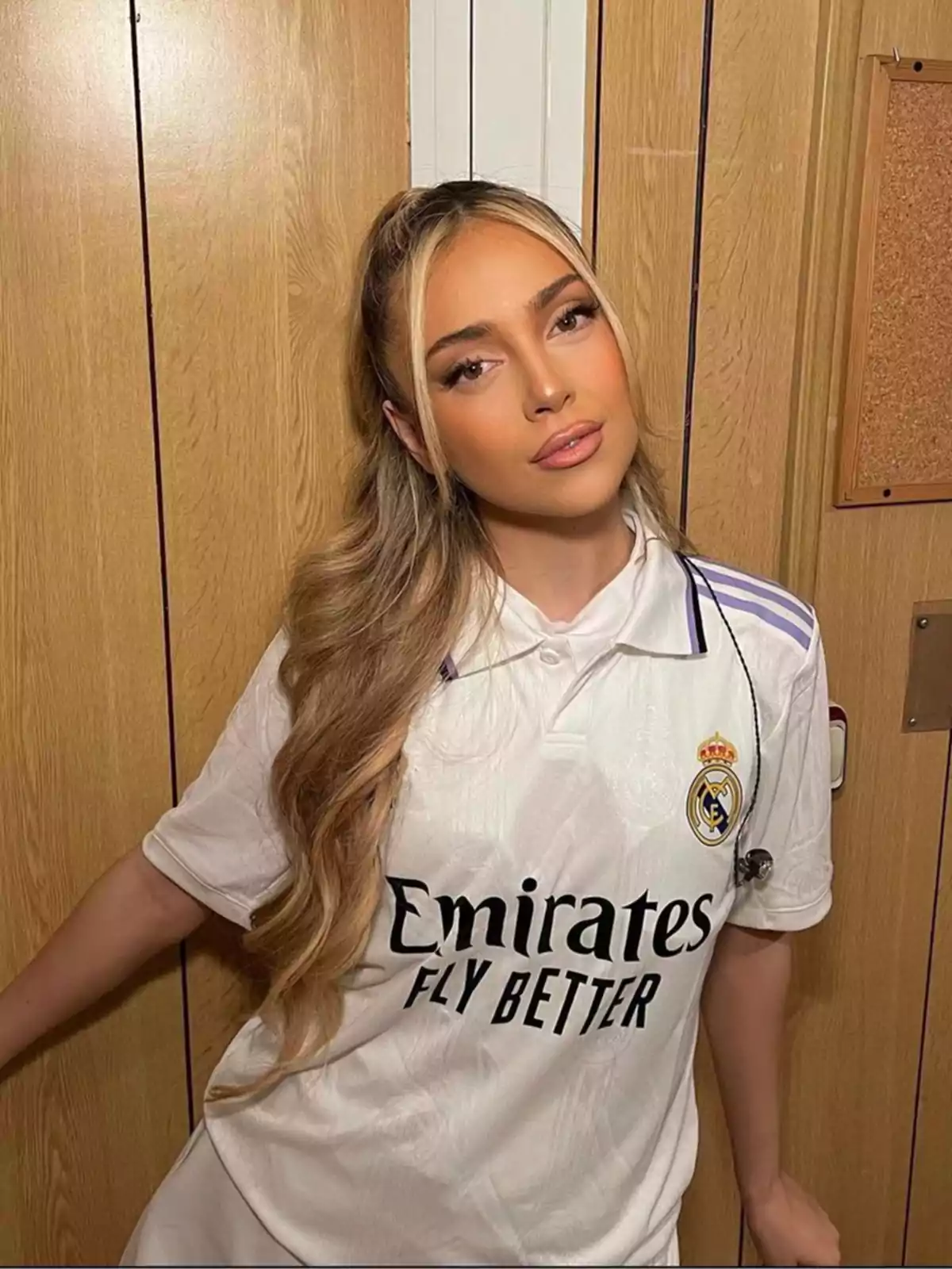 Una persona con cabello largo y rubio lleva puesta una camiseta blanca del Real Madrid mientras posa frente a una pared de madera.