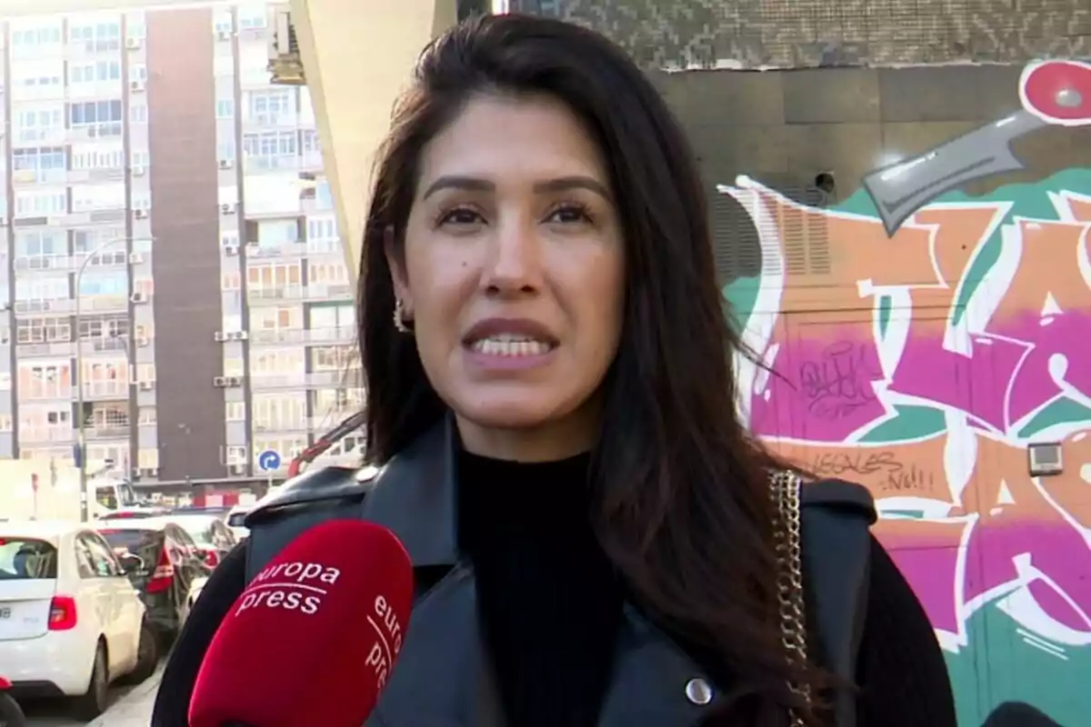 Gabriela Guillén con cabello oscuro hablando frente a un micrófono de Europa Press en una calle con grafitis y edificios de fondo.