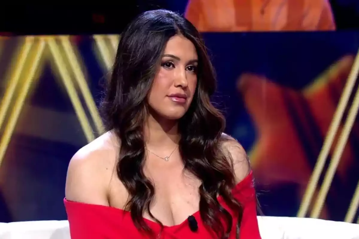 Una mujer con cabello largo y ondulado lleva un vestido rojo con hombros descubiertos mientras está sentada en un set de televisión con luces de fondo. Una mujer con cabello largo y ondulado lleva un vestido rojo con hombros descubiertos mientras está sentada en un set de televisión con luces de fondo.