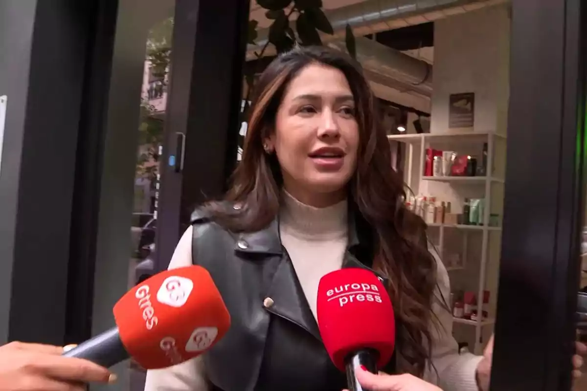 Una mujer con cabello largo y suelto está siendo entrevistada por dos micrófonos de medios de comunicación frente a un establecimiento con productos en estanterías. Una mujer con cabello largo y suelto está siendo entrevistada por dos micrófonos de medios de comunicación frente a un establecimiento con productos en estanterías.