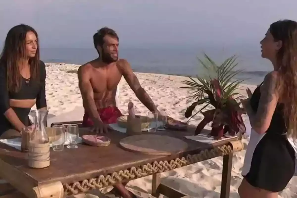 Tres personas conversan alrededor de una mesa en la playa con el mar de fondo. Tres personas conversan alrededor de una mesa en la playa con el mar de fondo.
