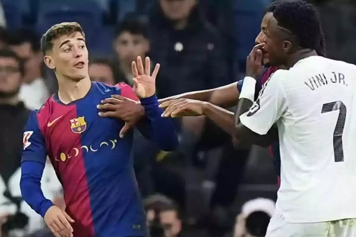 Dos jugadores de fútbol interactúan en el campo durante un partido, uno de ellos lleva la camiseta del Barcelona y el otro la del Real Madrid.