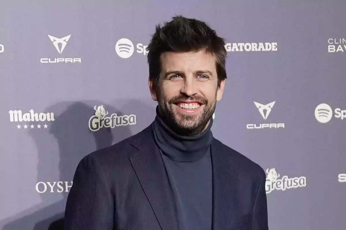 Un hombre sonriente con barba y cabello oscuro lleva un suéter de cuello alto y una chaqueta oscura, posando frente a un fondo con logotipos de marcas.