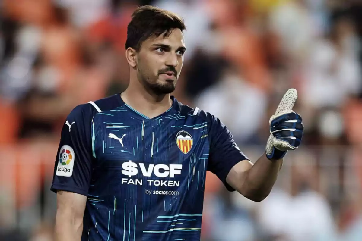 Un jugador de fútbol con uniforme azul oscuro del Valencia CF levanta el pulgar en señal de aprobación durante un partido. Un jugador de fútbol con uniforme azul oscuro del Valencia CF levanta el pulgar en señal de aprobación durante un partido.