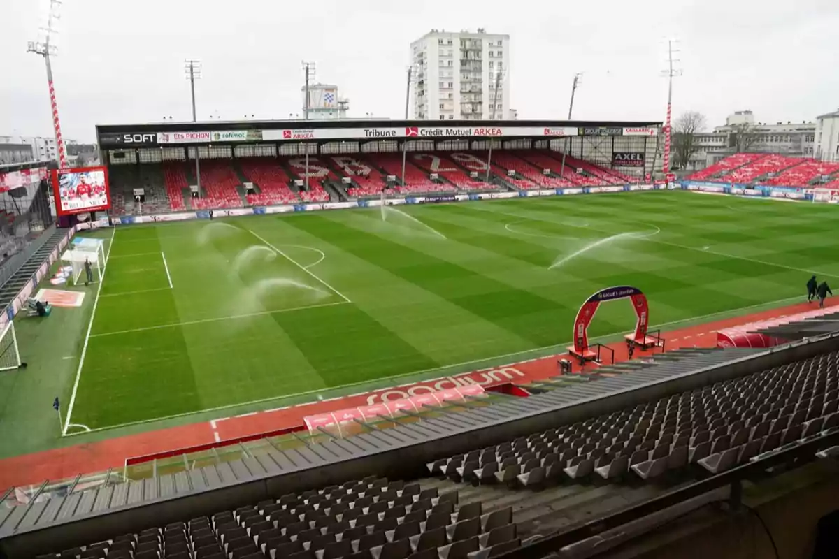 Un estadio de fútbol vacío con el césped siendo regado y las gradas rojas y grises.