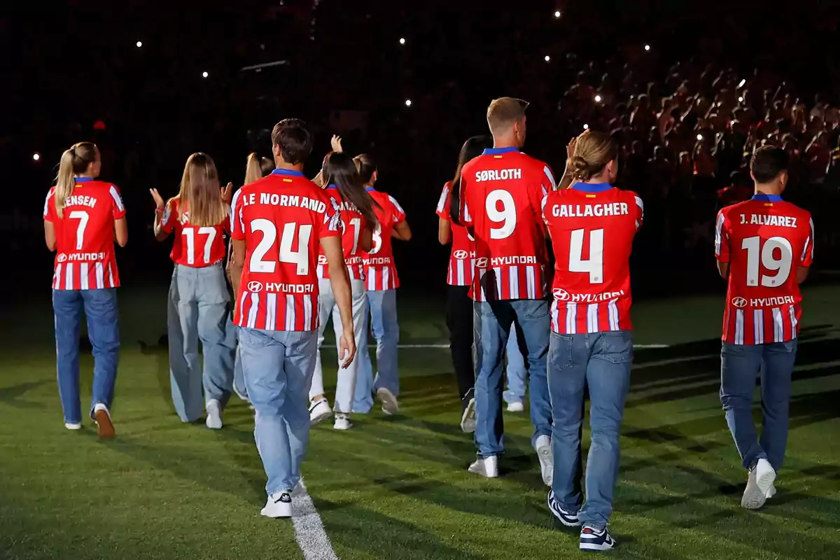 Jugadores de fútbol con camisetas rojas y blancas de un equipo, caminando sobre el césped mientras saludan a los aficionados en un estadio iluminado.