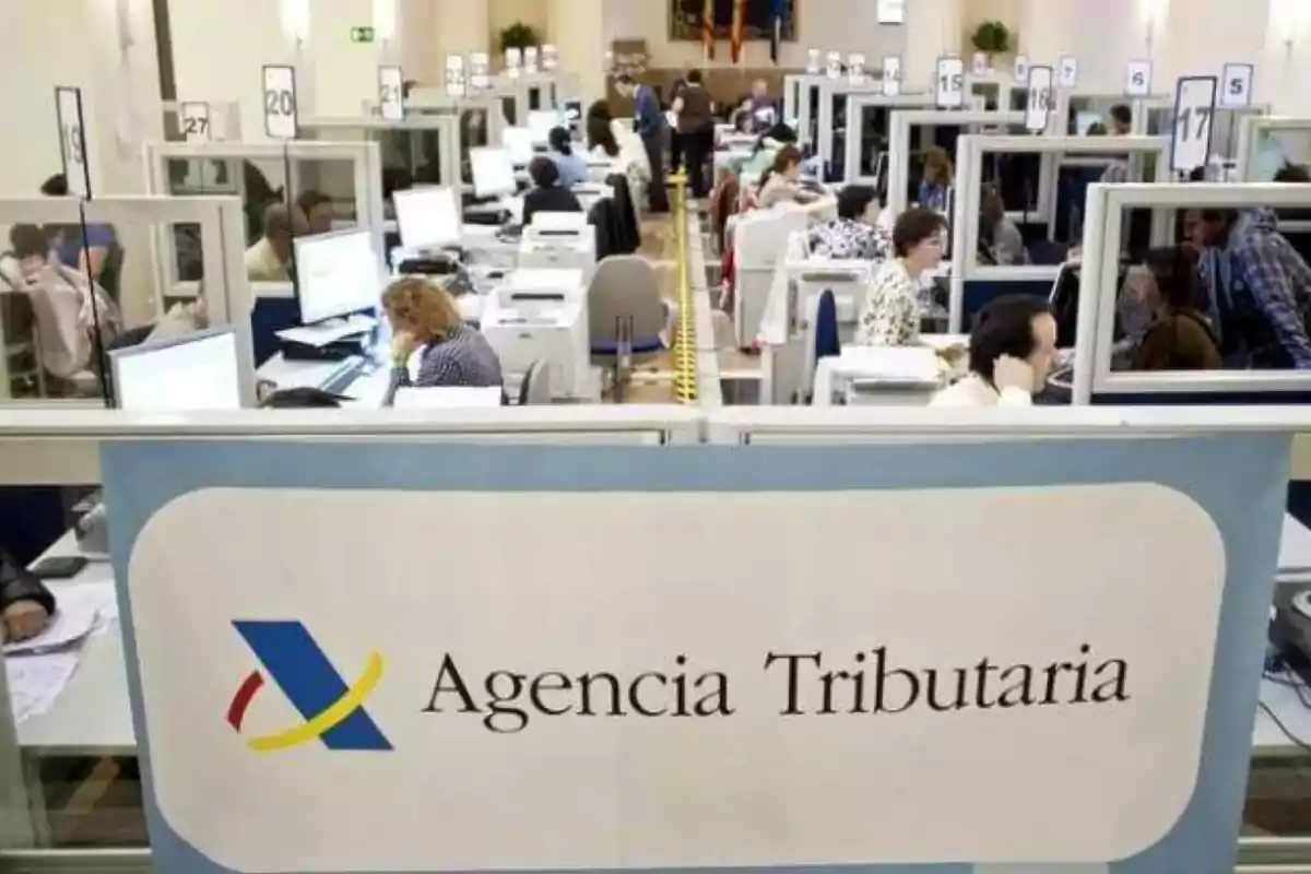 Oficina de la Agencia Tributaria con empleados trabajando en sus escritorios y atendiendo a personas.