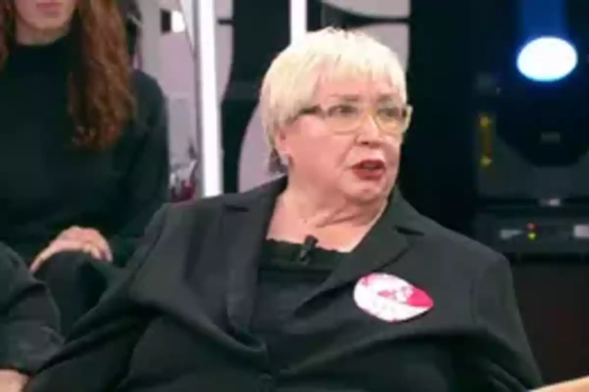 Una persona con cabello corto y canoso, usando gafas y un traje oscuro, está sentada en un estudio de televisión. Una persona con cabello corto y canoso, usando gafas y un traje oscuro, está sentada en un estudio de televisión.